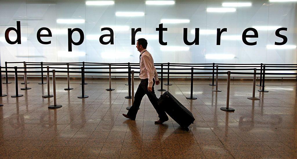 A man wearing black trousers and a white shirt pulling a suitcase in front of a giant sign saying departures that is brightly lit from behind