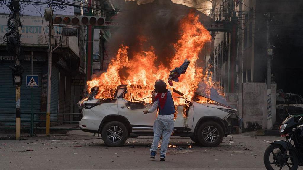 A person draped in Nepal's national flag throws an obejct towards a flaming vehicle in the street