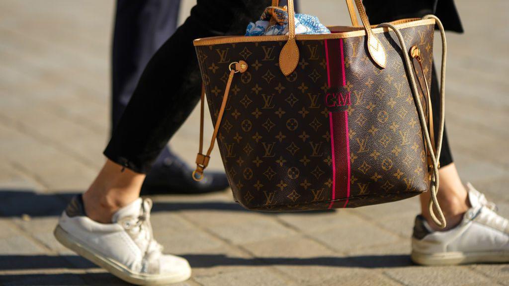 A guest wears black suede pants, a brown LV monogram in coated canvas Neverfull handbag from Louis Vuitton, white leather sneakers, outside Louis Vuitton, during Paris Fashion Week - Womenswear Spring/Summer 2023, on October 04, 2022 in Paris, France.