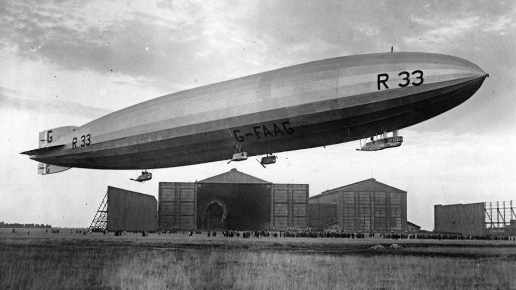 An airship in a black and white photograph. It hovers over a crowd of hundreds of people and in front of large air hangars. At the front of the ship it says "R33" in large black letters.