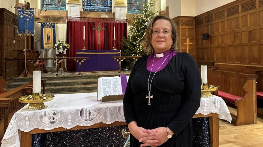 Rt Rev Tricia Hillas wearing a dog collar with a purple shirt under a black dress. There is a crucifix around hr neck and she is standing in front of a nativity scene in front of the alter in St George's Church in Douglas. There is a large Christmas tree behind her.