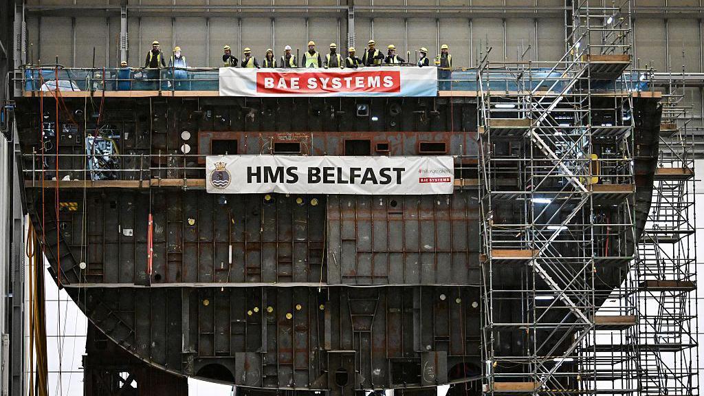 A line of people wearing hi-vis vest and hard hats, look very small compared to the structure of the HMS Belfast ship they are standing on. The vessel is still in construction so lots of scaffolding is visible.