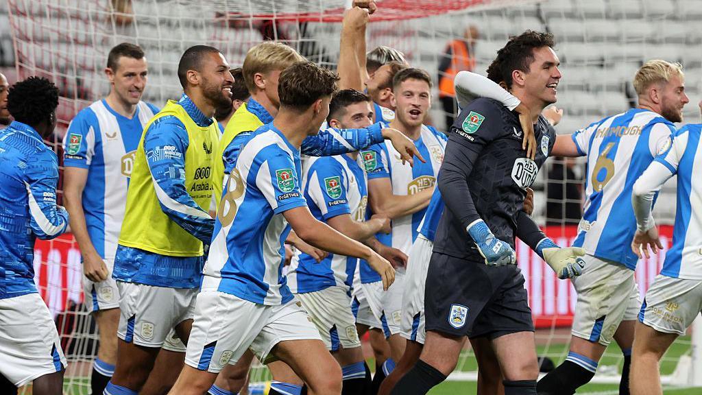 The Huddersfield Town players surround goalkeeper Lee Nicholls after his save from Milan Aleksic in the shootout