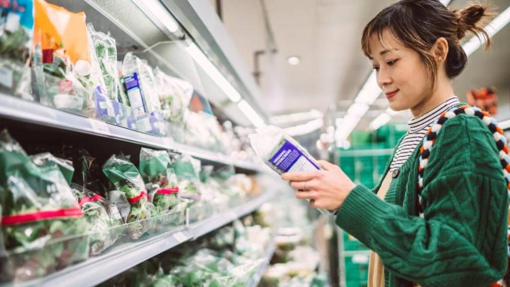 Woman inspects a bag of lettuce in the middle of a grocery aisle that primarily contains leafy greens.