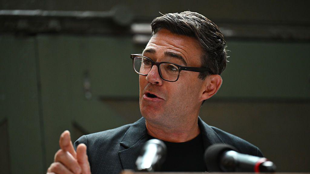 Andy Burnham making a speech at the People's History Museum in Manchester on Monday. He is gesturing with his right hand and is mid-speech. He is wearing glasses, a grey blazer and black shirt.