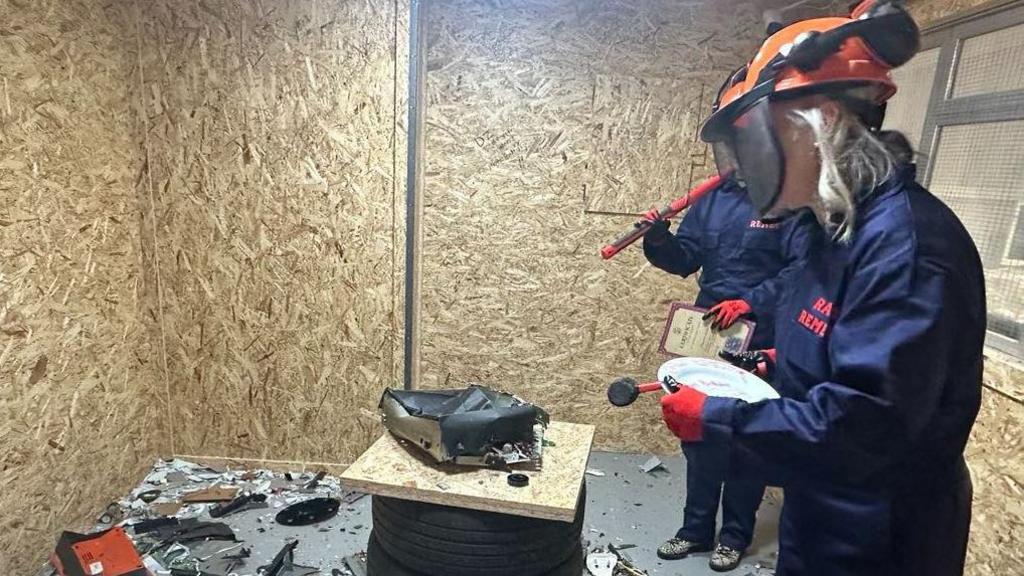 Claire Seward in the foreground dressed in mauve overalls and wearing an orange helmet and holding a mallet in a room covered in chipboard with a smashed up small appliance in the centre 