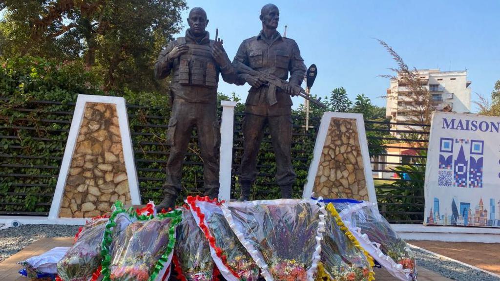 Full bronze statues in the likeness of the late Wagner private mercenary group chief Yevgeny Prigozhin (L), and his right-hand man Dmitru Utkin (R). Bouquets of flowers placed in the foreground and a green live fence in the background