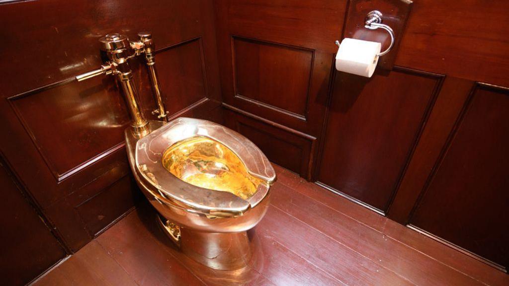 The gold toilet - a basin, seat and pipes - sitting in a corner of a room panelled with dark wood. There is a white toilet roll on a silver toilet roll holder.