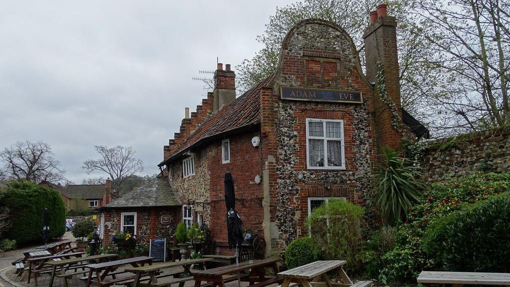The Dutch gable end of a red brick and flint pub, with the sign Adam and Eve above its second floor window. There are pub benches outside.