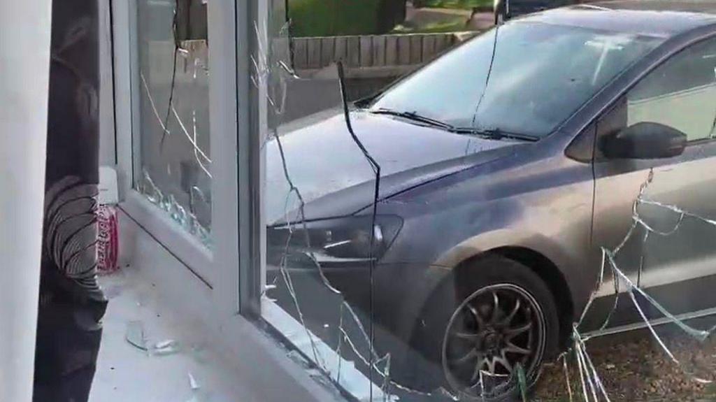 View from inside a house looking out through a shattered front window. The glass pane is covered in large cracks and several sections are broken, with shards of glass scattered across the white windowsill. Outside, a grey car is parked close to the window on a residential street lined with trees and hedges. The image appears to document damage to the property, with the broken window as the main focus.