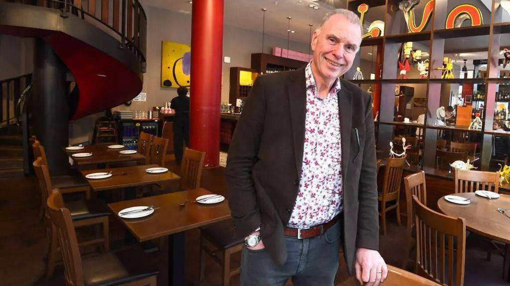 Peter Kinsella standing in a Spanish restaurant he owns in Liverpool. He is wearing a floral shirt, with jeans and a dark brown jacket