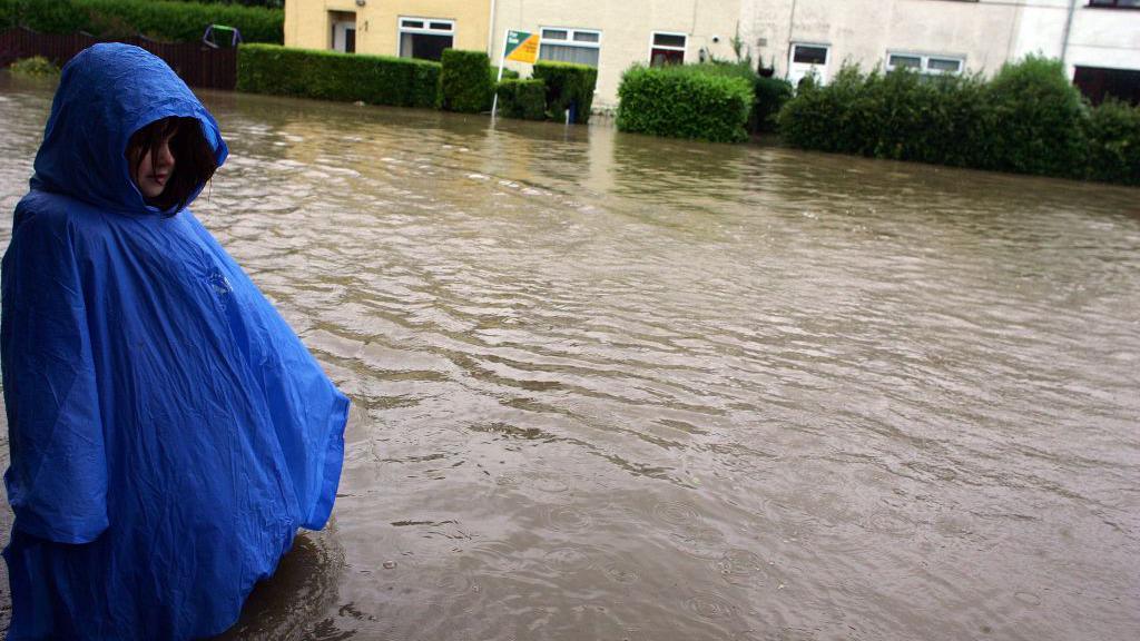 A girl in a bright blue poncho standing in flood water up to her waist in Hull in 2007. The road is covered in water which extends into the gardens of a row of houses at the top of the picture.