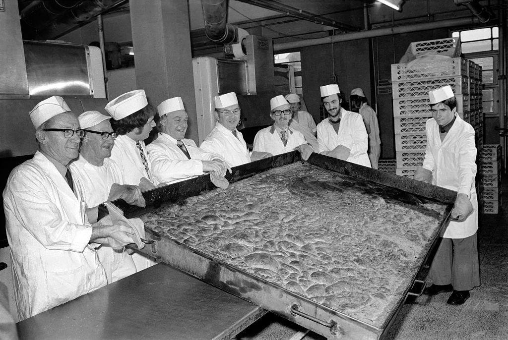 an archive photograph of eight men hold a massive tray while wearing oven gloves with a roasted yorkshire pudding filling the whole tray while standing in an industrial kitchen