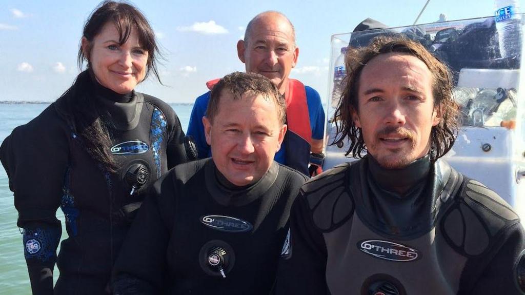 A woman and two men in diving suits with a third man behind in a boat in the Thames Estuary in 2014