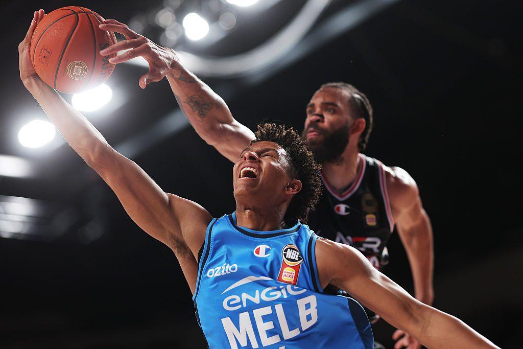 A player in a blue jersey stretches toward the hoop for a layup while a defender reaches over from behind, creating a tense, high-energy moment of contact under bright arena lights.