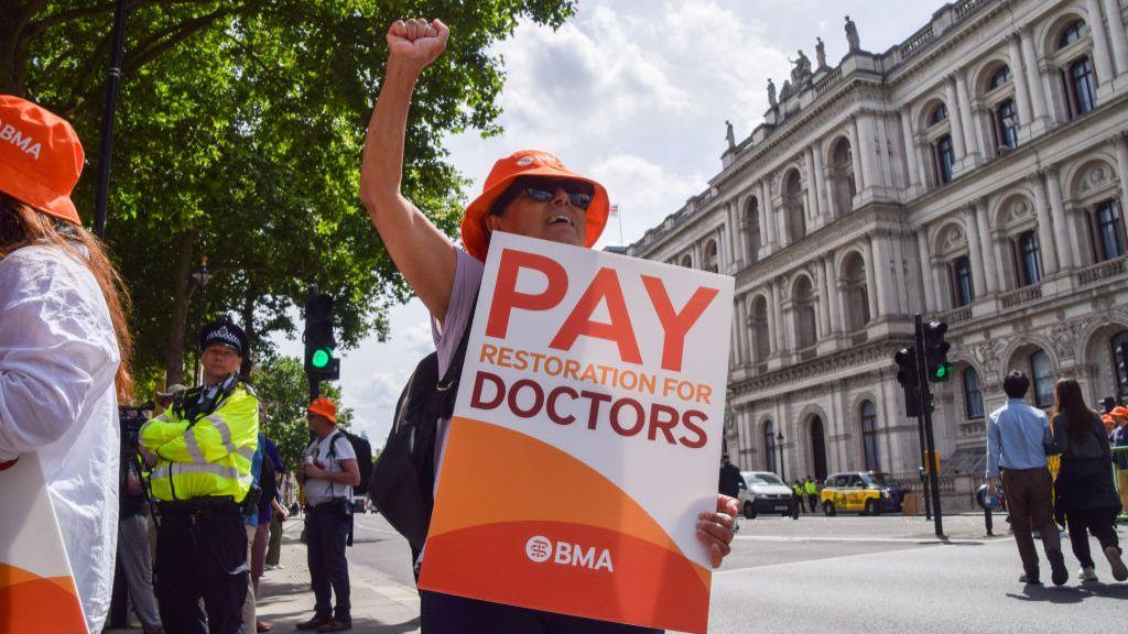 A junior doctor holds a placard that says "Pay restoration for doctors" during a rally in London in June last year. They are holding up one arm and wearing an orange British Medical Association hat and sunglasses, while their body is largely obscured by their sign. Behind them other protesters, a police officer and pedestrians are standing around.