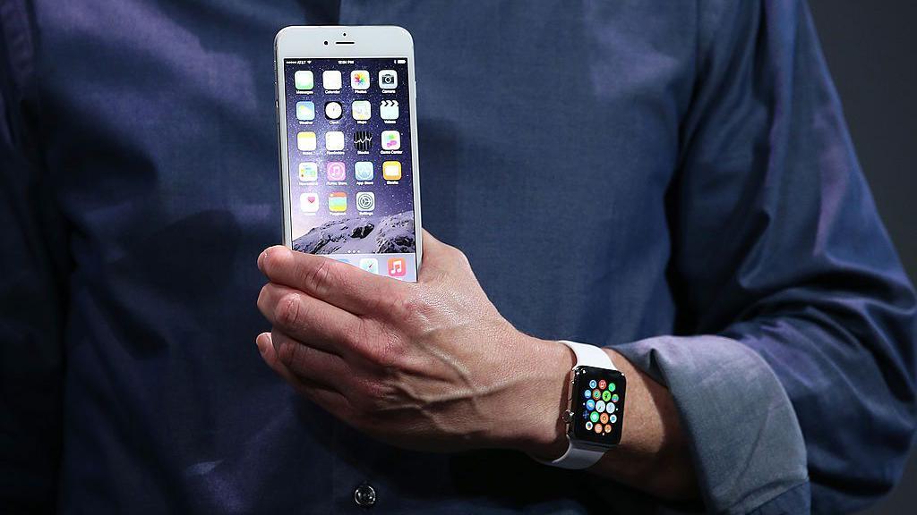 Close up image of a man's hand holding a white iPhone 6, with an Apple Watch on his wrist. His dark blue shirt is rolled up at the cuff.