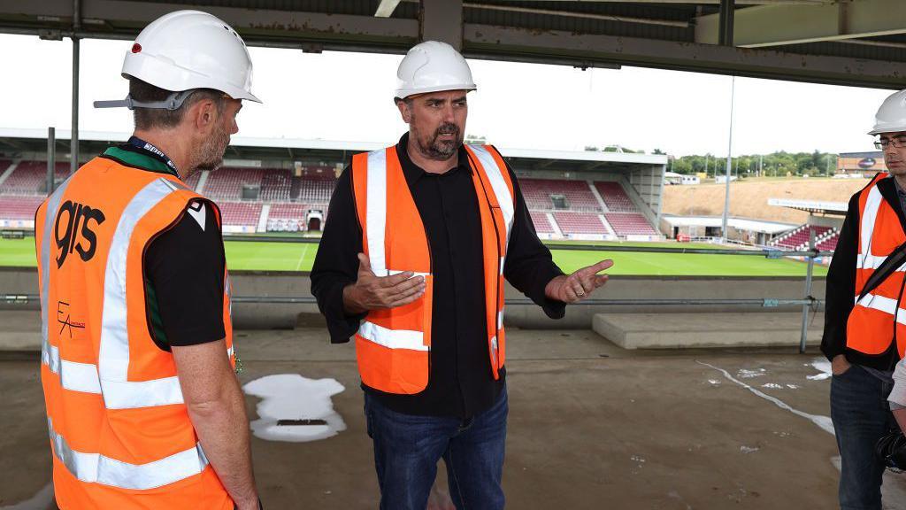 A man in a black top, blue jeans, a orange and white high visability jacket and a white hard hat speaks to several people dressed the same, in the construction site of the East Stand at Northampton Town Football club. The west stand and the pitch can be seen in the background. 