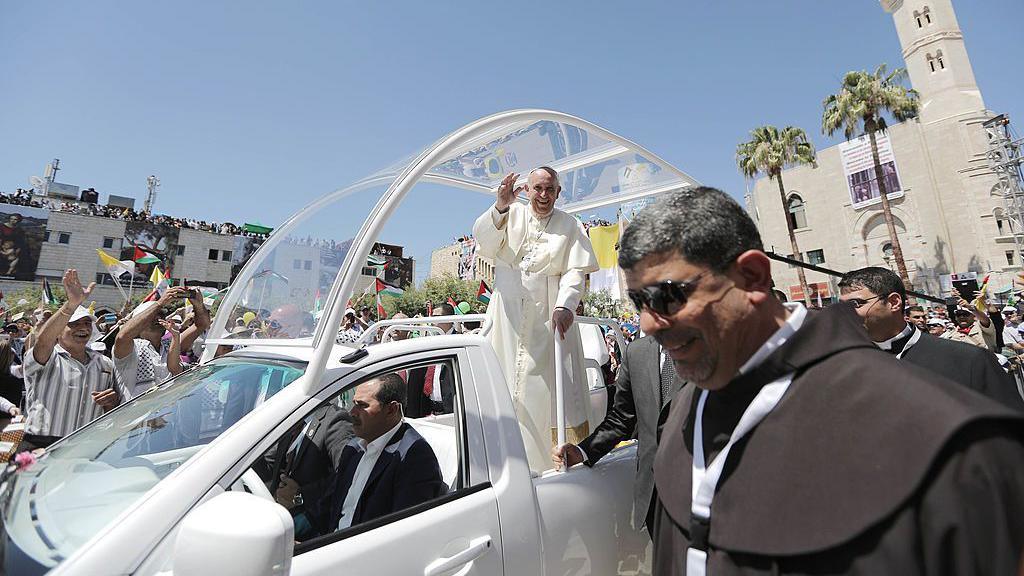 The pope, waving to people, stands in the white popemobile, smiling