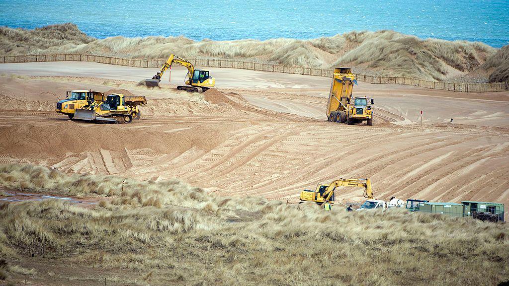Four heavy diggers and transporters - all yellow - work at clearing away land by the sea and dunes. The sandy ground has been rendered flat and featureless. 