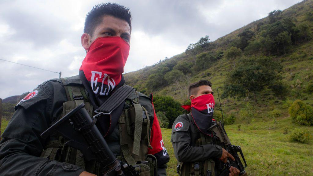 Two fighters of the ELN with red and black face coverings and rifles in a forest clearing in the mountains of Colombia in 2025