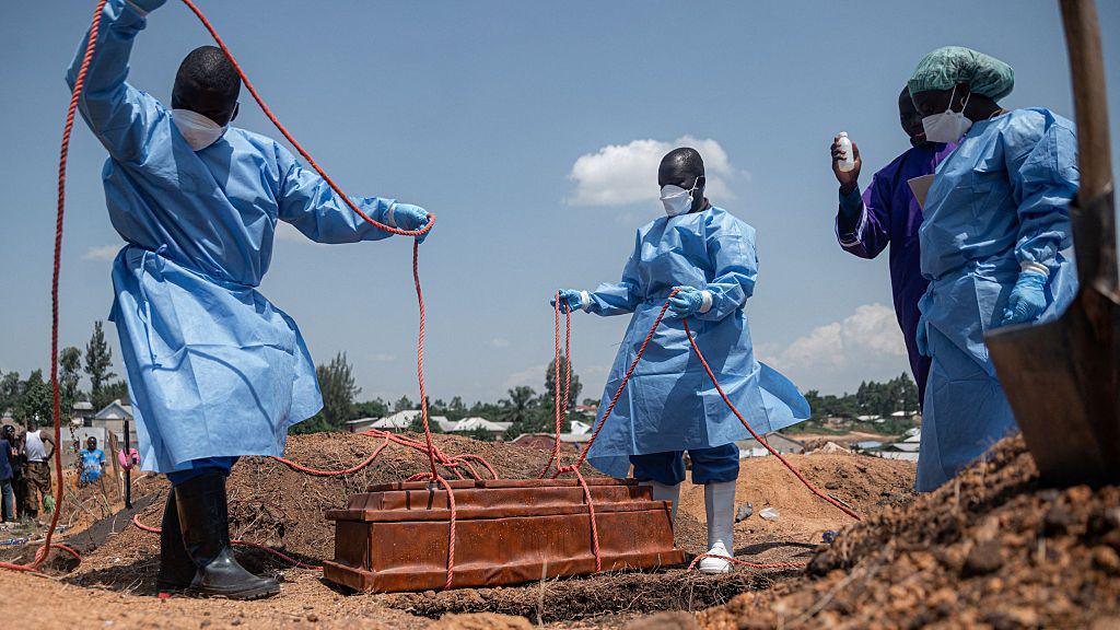 Four people wearing personal protective gear lower a coffin into a grave using ropes