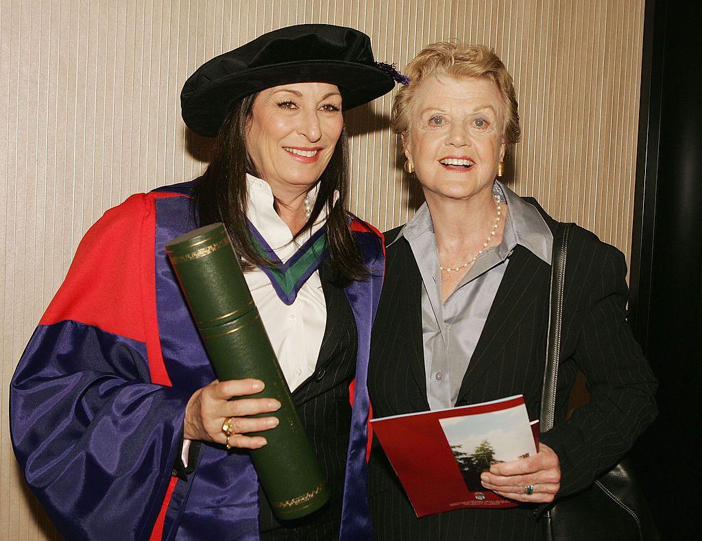 A smiling woman (Huston) with long black hair, wearing a graduation cap, and purple and red gown, holds a green scroll. She looks off camera. Shes closely standing beside another woman (Lansbury), who is wearing a grey shirt, black jacket, and has short blonde hair. She holds a pamphlet in her hand.  