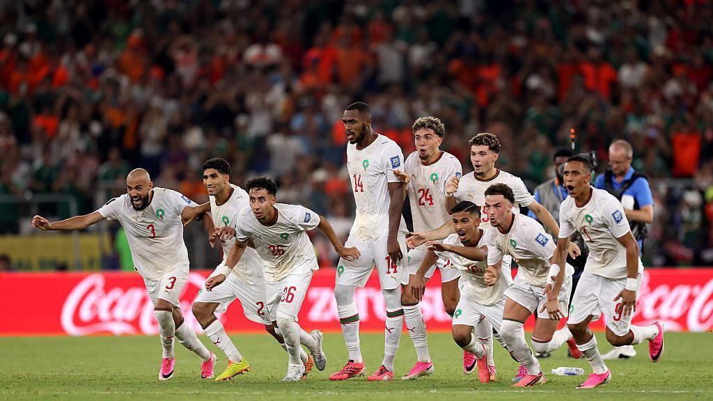 Morocco players linked in arms on the halfway line sprint towards their goalkeeper after winning the penalty shootout