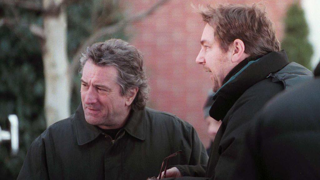 Robert De Niro, with feathered hair and a dark jacket, stands outdoorsnext to another man, who is holding a pair of glasses and wearing a black jacket.