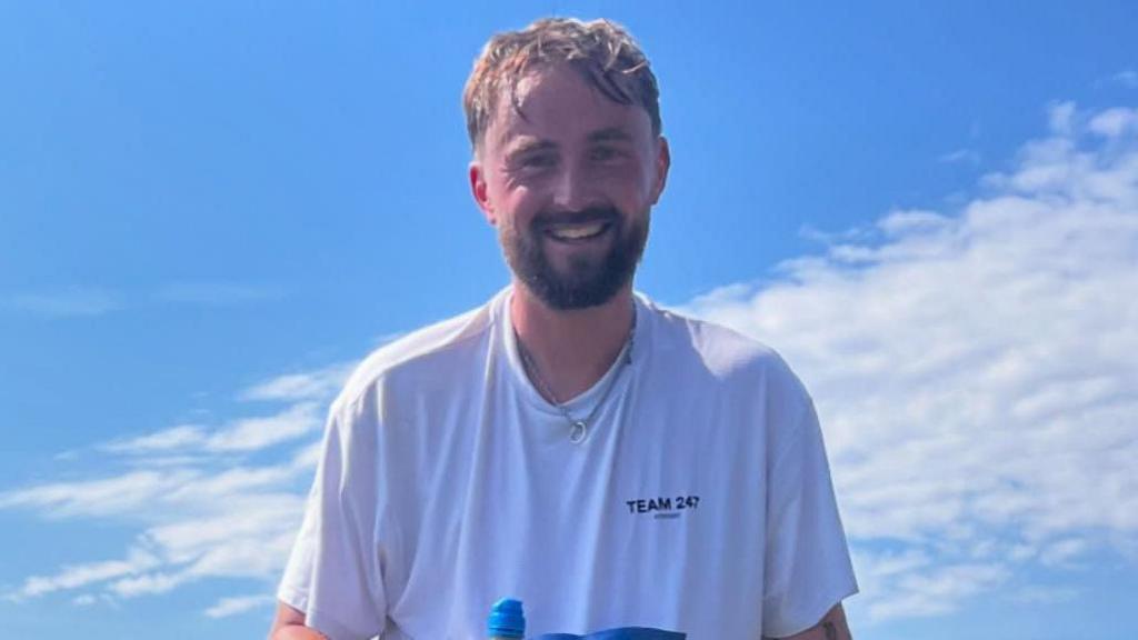 Ryan Cruikshanks has short brown hair and a short brown beard. He stands in front of the blue sky with a cloud across it. He wears a white top with a "Half DRAM 2025" label and holds a bottle of Lucozade.