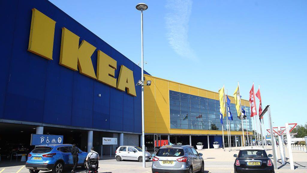 An image of an Ikea store. It is a large blue and yellow-coloured building with the word "IKEA" in large yellow lettering. There are flags on poles and cars parked in front of the store.