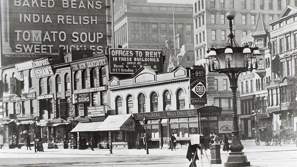 Historic black and white photo showing an ornate streetlamp in front of buildings and old billboards