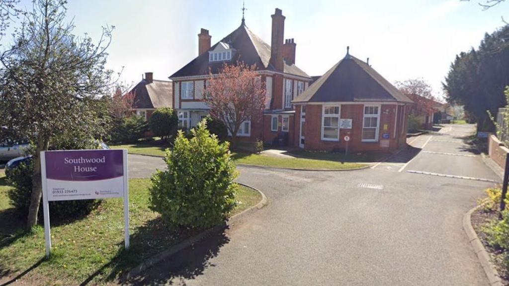 A red brick care home in the background. A grey driveway leads up to it and and there is a white sign in the foreground which states Southwood House.