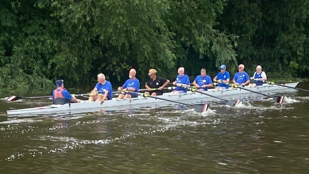 A group of eight men in blue t-shirts rowing. There is a cox at the back of the boat.