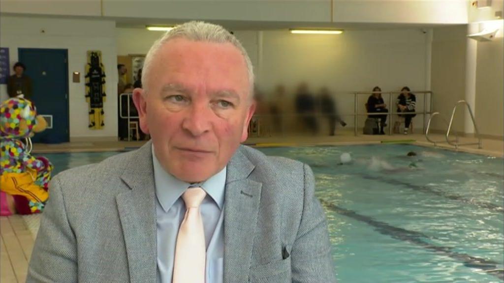 Gary Gentle is sitting in front of a swimming pool, where there are children swimming. He has white hair and is wearing a grey suit jacket, blue shirt and salmon coloured tie.