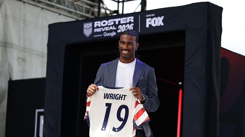 Haji Wright wearing a blue suit jacket and white t-shirt poses with his USA shirt at the event to reveal the USA's World Cup squad