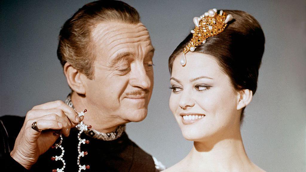 David Niven looking at Claudia Cardinale while holding up jewellery 