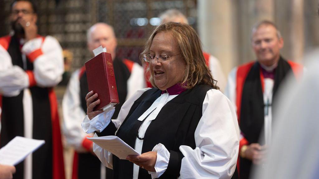 The Venerable Tricia Hillas holding up a bible and taking an oath. Other members of the clergy look on in the background.