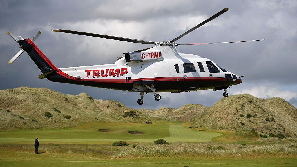 A large white, black and red liveried helicopter with TRUMP branding on the fuselage is coming in to land on the fairway at one of the links courses. Trump, in a dark blue suit, can be seen on the edge of frame, standing waiting for it to land. The sky is grey and filled with clouds. 