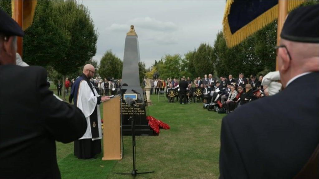 A vicar stands next to a black granite memorial, topped with a golden sphinx, at an outdoor ceremony as people gather around