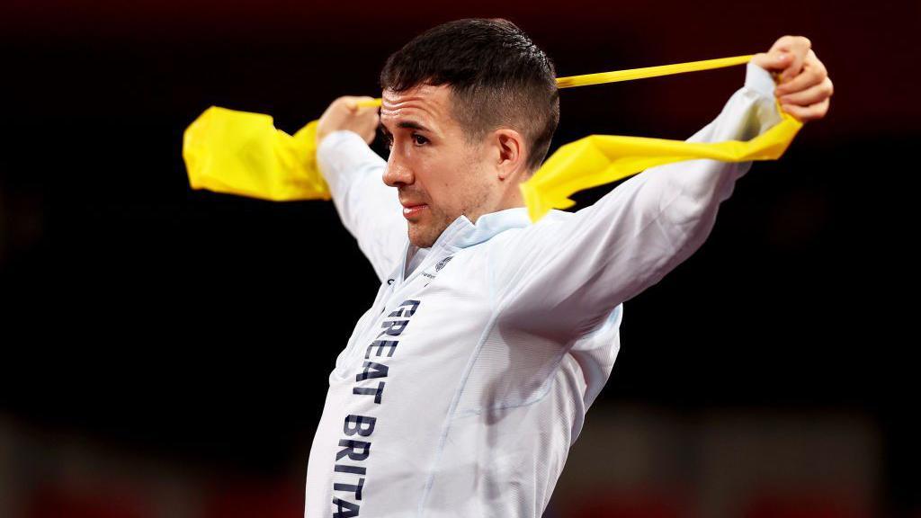 A man in a white sports jacket stretches his arms wide behind his head with a yellow band between his hands. 