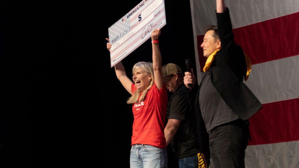 Elon Musk handing a woman a giant cheque on 21 October