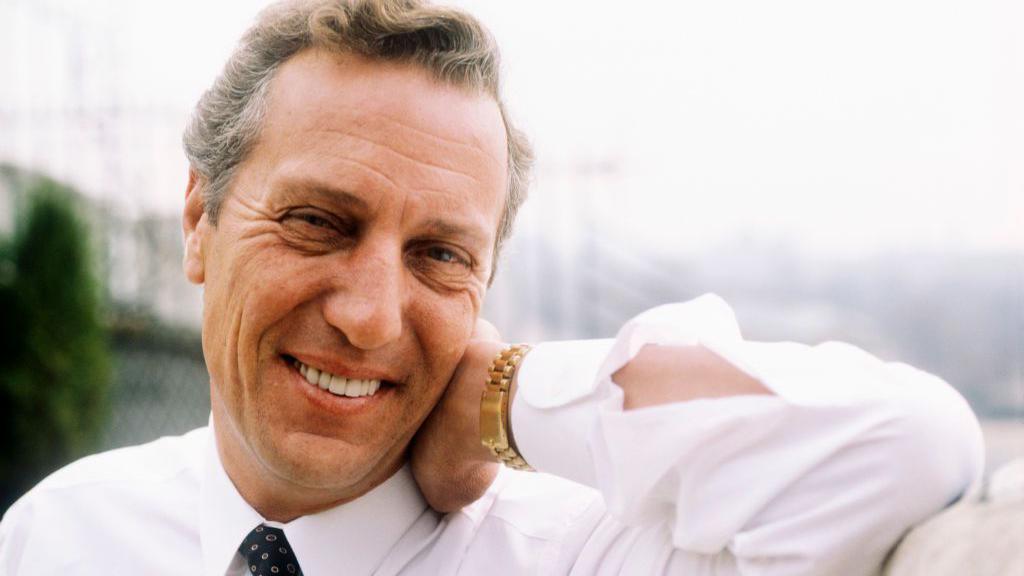 Frederick Forsyth photographed in the 1980s. He is smiling at the camera and his his hand on his neck.