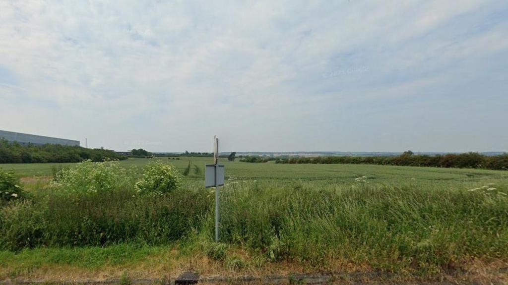 A field with tall grass in the foreground and shorter grass beyond. There is a hedge in the background. There is a low-level warehouse to the left and a road sign in the centre of the picture.