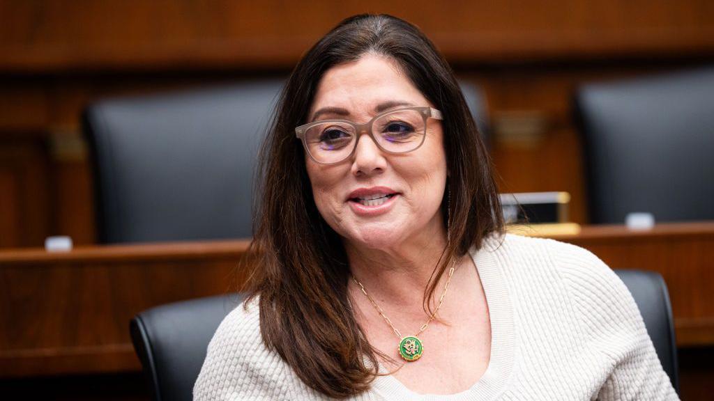 Lori Chavez-DeRemer is seen seated in a congressional hearing room wearing glasses and a beige shirt. 