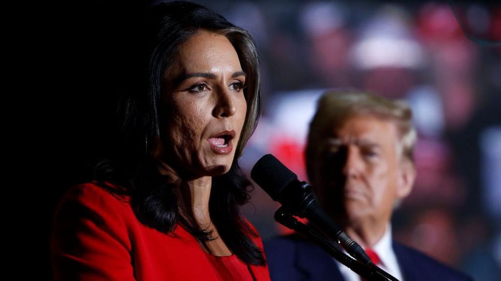 Tulsi Gabbard, wearing a red pantsuit, speaks into a microphone. Trump's head is blurry in the background