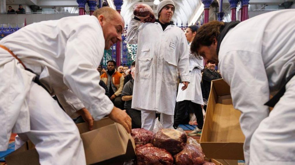 Butchers wearing white coats handle large lumps of meat while a seated audience looks on.