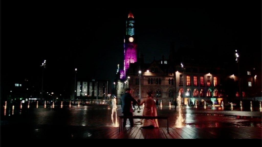 A scene from Virdee with the two protagonists in wedding outfits in front of the mirror pool and City Hall at night