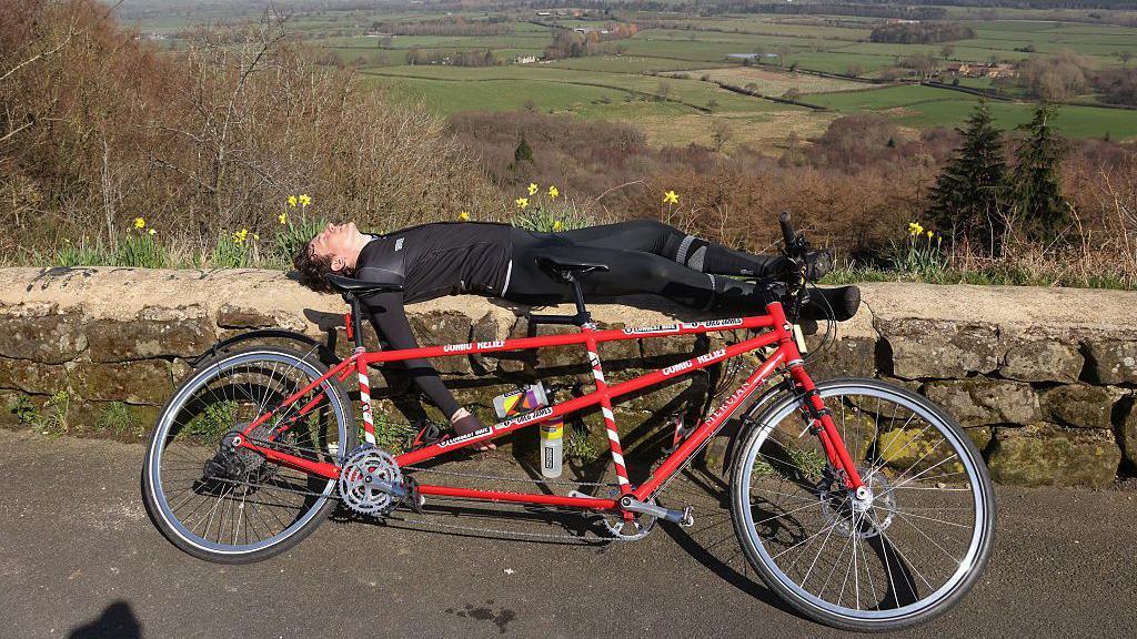 Radio presenter Greg James lying down on a stone wall in the countryside next to a tandem bike.