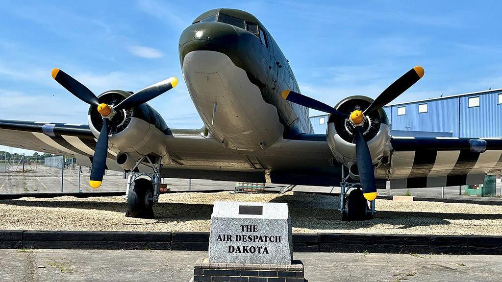The restored Dakota Gate Guardian at RAF Brize Norton 47 Air Despatch Royal Logistic Corps in Oxfordshire on a sunny day. There is a marble badge and its name in front of the plane.